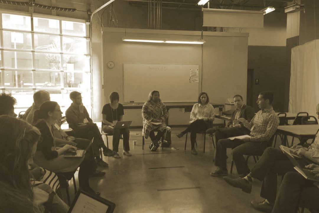 A group of people sitting in chairs arranged in a circle in a large room with a glass-paneled wall in the background.